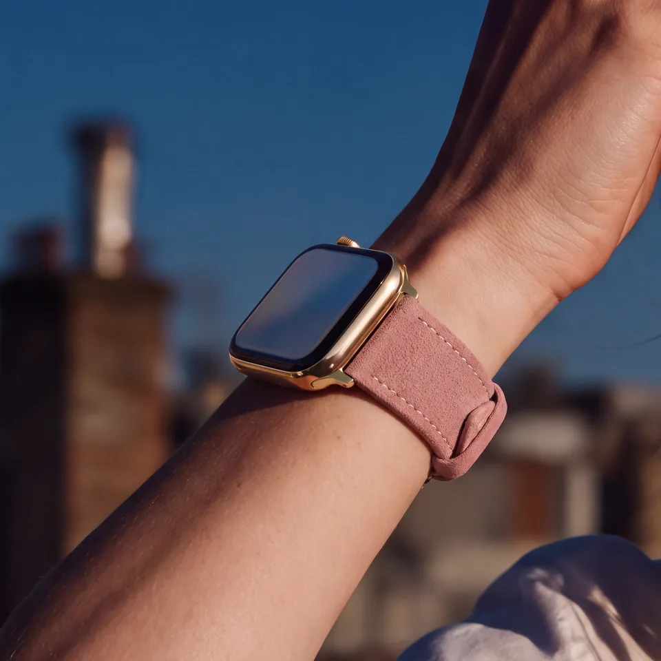 Nahaufnahme eines Handgelenks mit goldener Apple Watch und rosé Alcantara-Armband von INDISTRAPS vor Dächern im Abendlicht