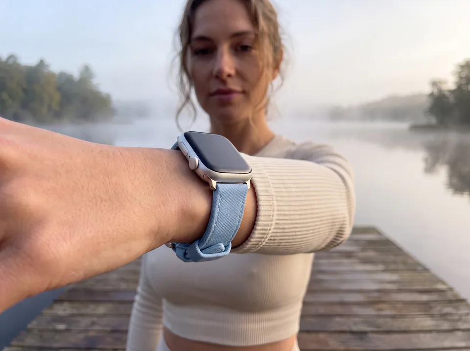 Frau auf einem Holzsteg am nebligen See streckt den Arm aus und trägt eine Apple Watch mit hellblauem Canvas-Armband von INDISTRAPS – Variante 1