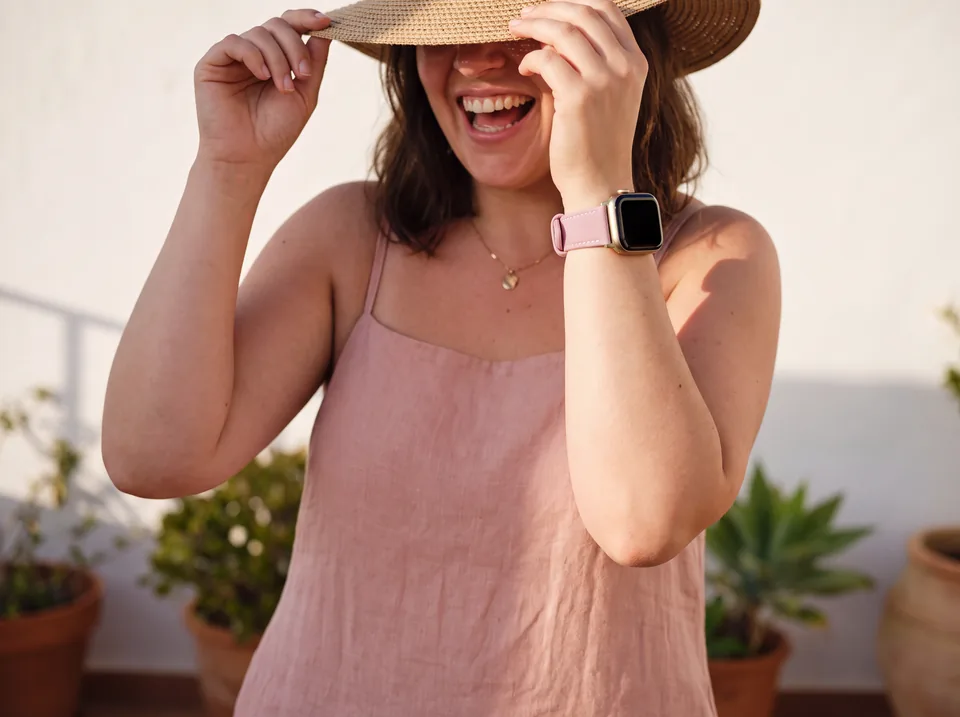 Lachende Frau mit Strohhut und rosa Leinentop auf einer Terrasse trägt eine Apple Watch mit altrosa Alcantara-Armband von INDISTRAPS – Variante 1