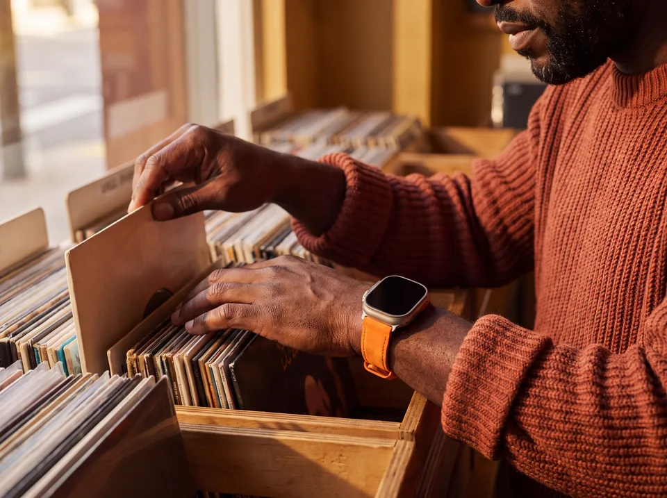 Mann in rostrotem Strickpullover stöbert im Plattenladen durch Vinyl und trägt eine Apple Watch Ultra mit orangefarbenem Canvas-Armband von INDISTRAPS – Variante 1
