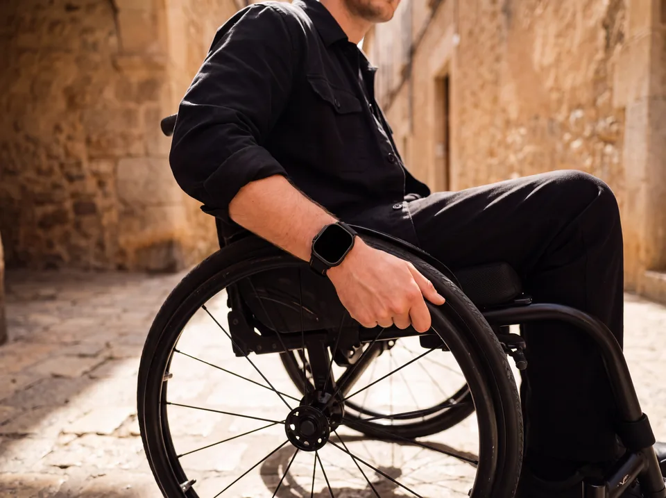 Mann im Rollstuhl in schwarzem Outfit in einer sonnigen Altstadtgasse trägt eine Apple Watch mit schwarzem Alcantara-Armband von INDISTRAPS – Variante 1