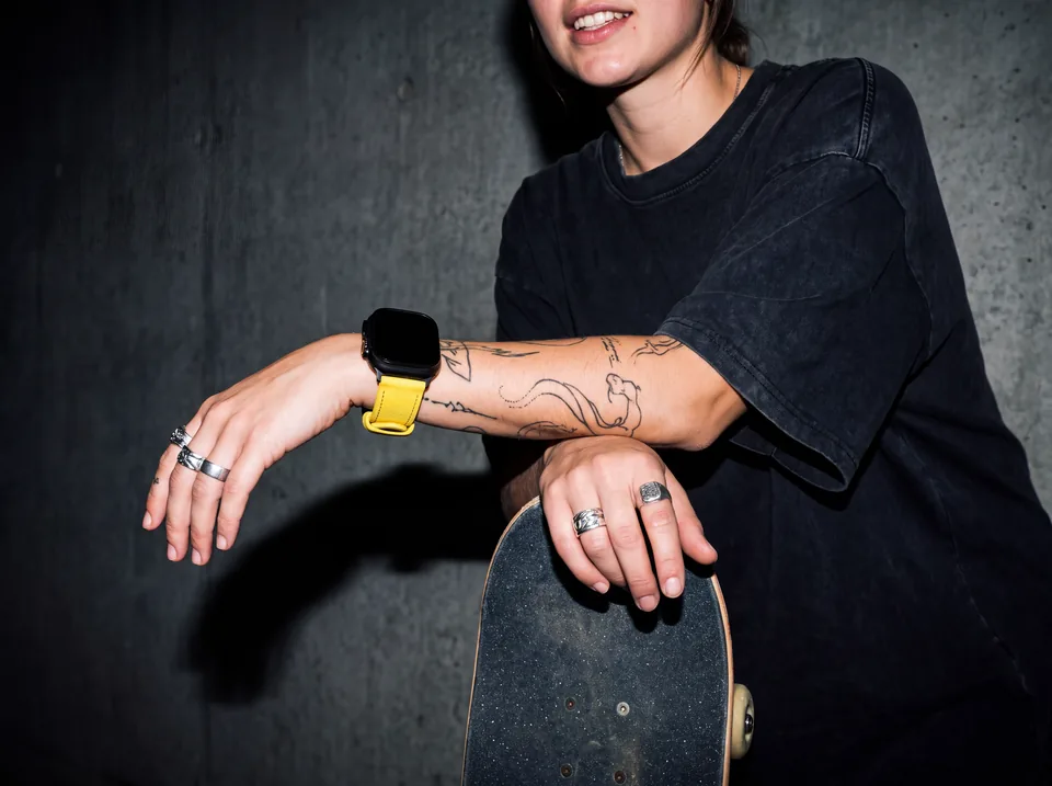 Skaterin mit Tattoos und Ringen lehnt auf ihrem Skateboard vor Betonwand und trägt eine Apple Watch mit gelbem Alcantara-Armband von INDISTRAPS – Variante 1
