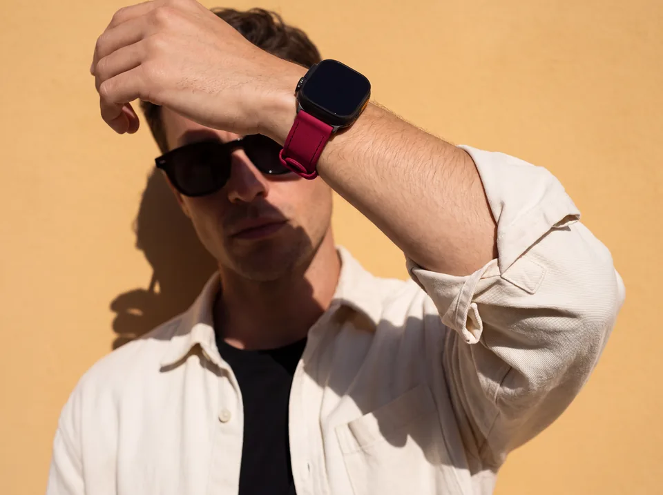 Mann in cremefarbenem Overshirt und Sonnenbrille vor gelber Wand im harten Sonnenlicht trägt eine Apple Watch mit kirschrotem Alcantara-Armband von INDISTRAPS – Variante 1